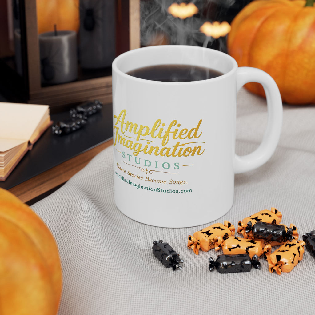 White mug with 'Amplified Imagination Studios' text, steaming coffee, Halloween-themed cookies, and pumpkins on a table.